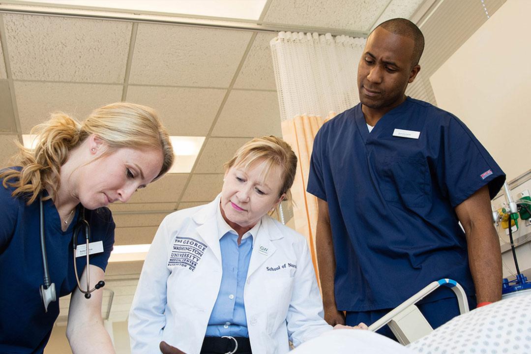 Two nurses in scrubs and a doctor in a GW lab coat work around a patient's bed